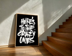 Framed poster with 'Here's to the crazy ones' text on a wall next to wooden stairs.