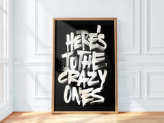 Framed poster with 'Here's to the crazy ones' text on a black background in a room with white walls and wooden floor.