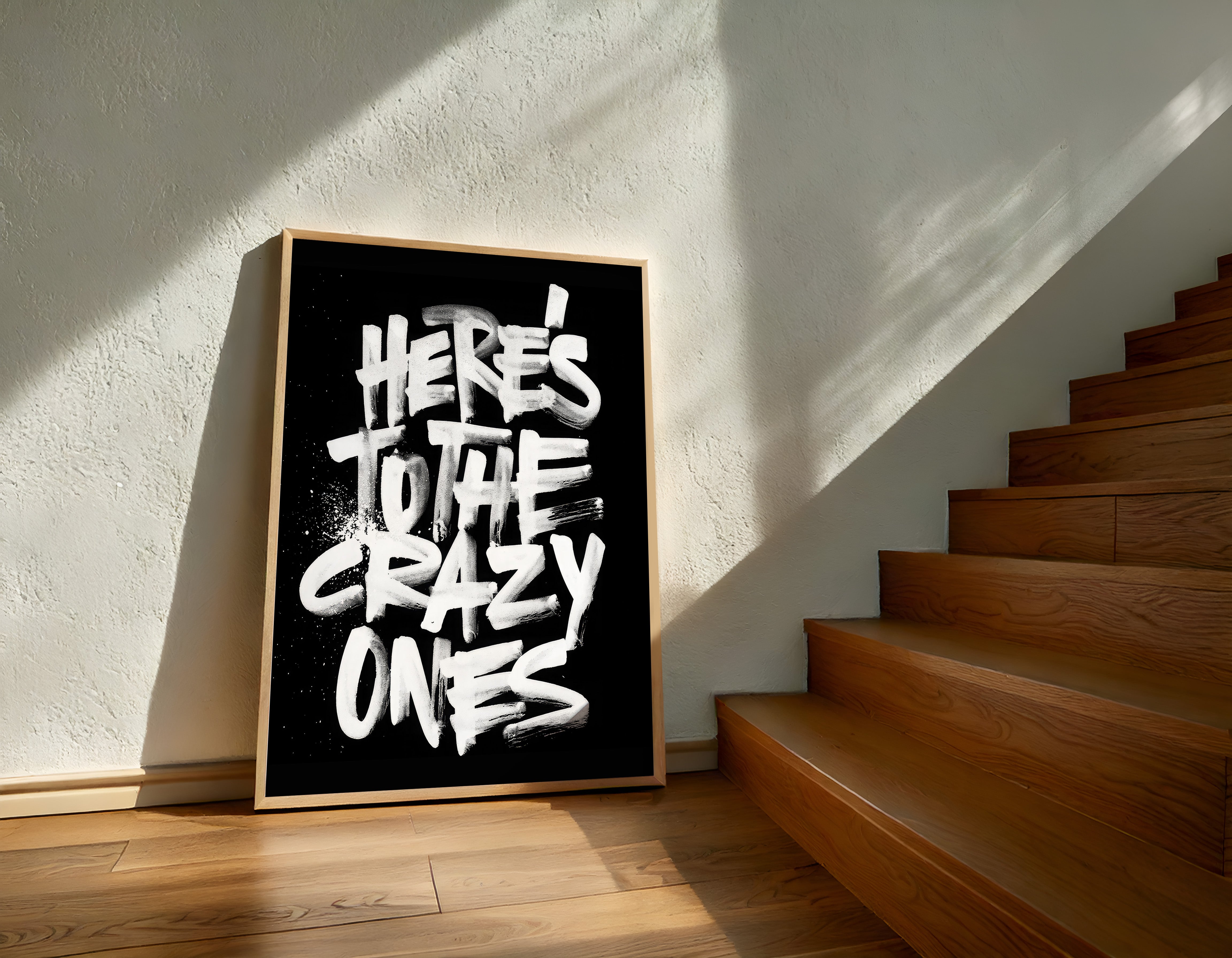 Framed poster with 'Here's to the crazy ones' text on a wall next to wooden stairs.